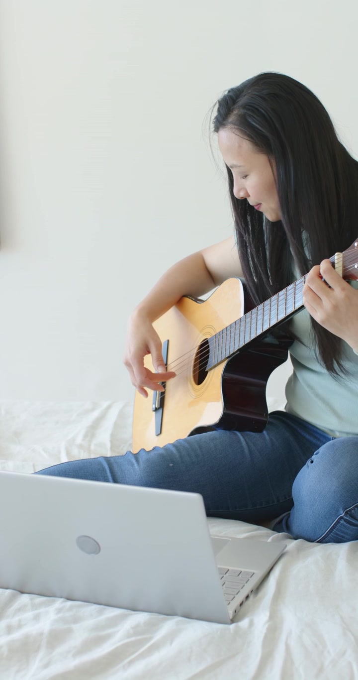 Video vertical: Mujer siguiendo una lección en el portátil, punteando y rasgueando la guitarra en la cama para practicar