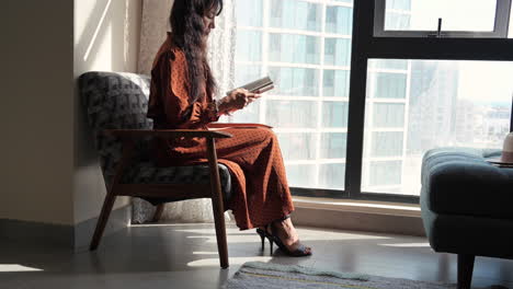 Mujer-Sentada-En-Una-Silla-Leyendo-Un-Libro-Junto-A-La-Ventana-De-Cristal-De-La-Sala-De-Estar-Con-Fondo-De-Luz-Solar-Matutina,-Bahréin