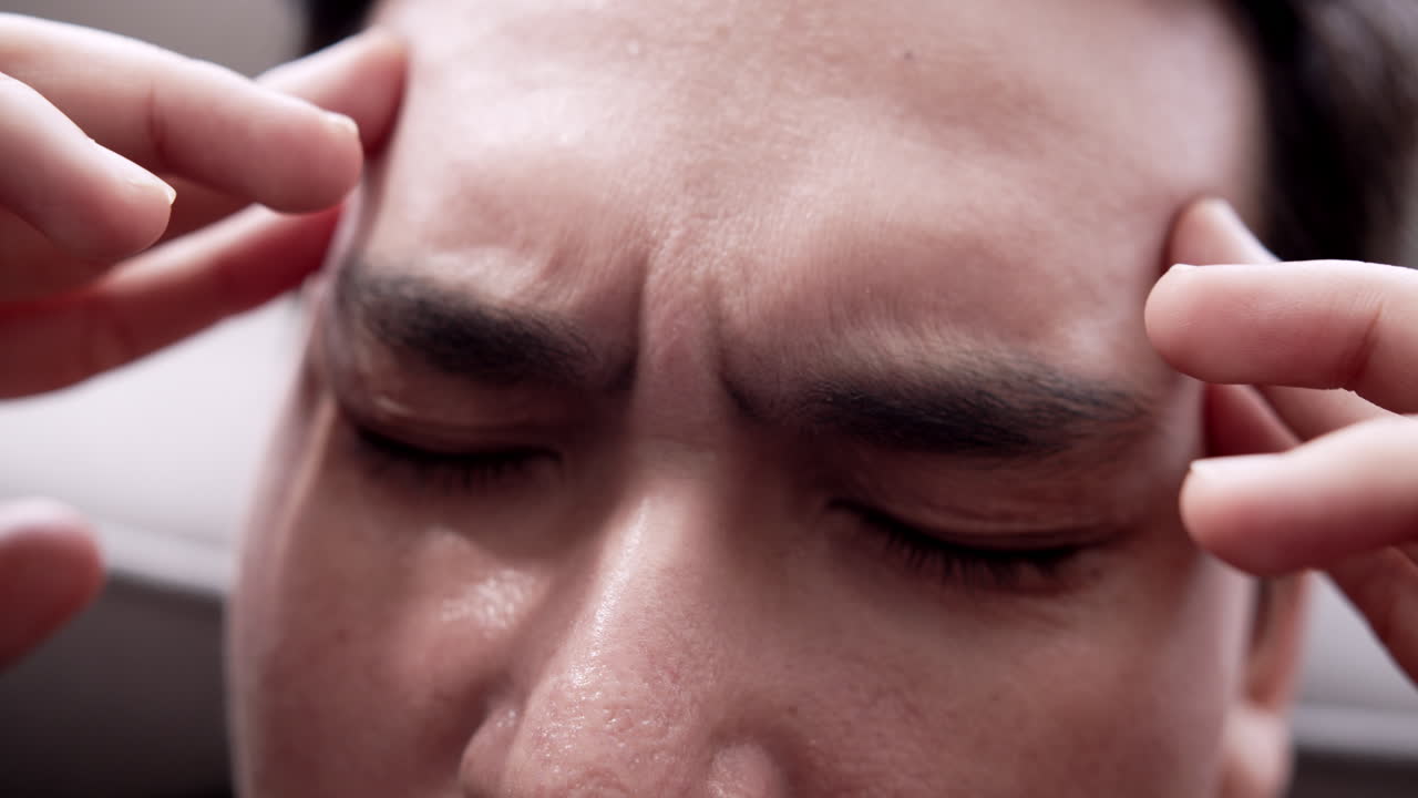 Close up of an Asian man massaging his head, looking anxious about the problem he is facing. Mood of sadness, depression, and disappointment.