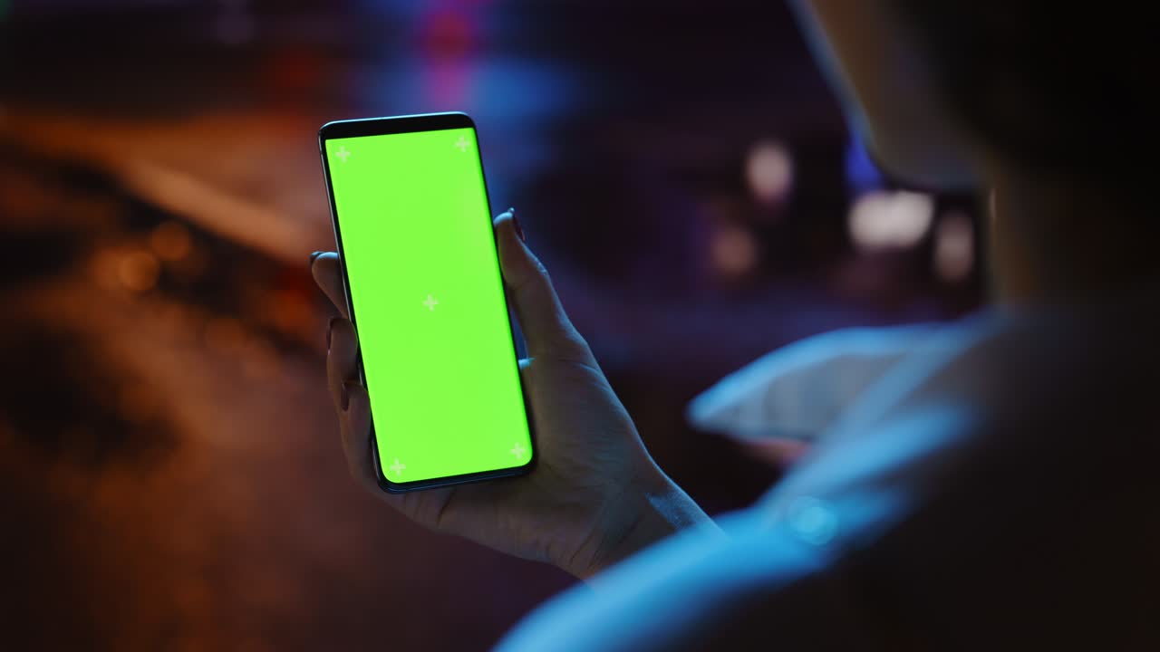Beautiful Woman using Chroma Key Smartphone while Walking Through Night City Street Full of Neon Light. Female Using Green Screen Mobile Phone. Over the Shoulder Static Medium Close-up Shot