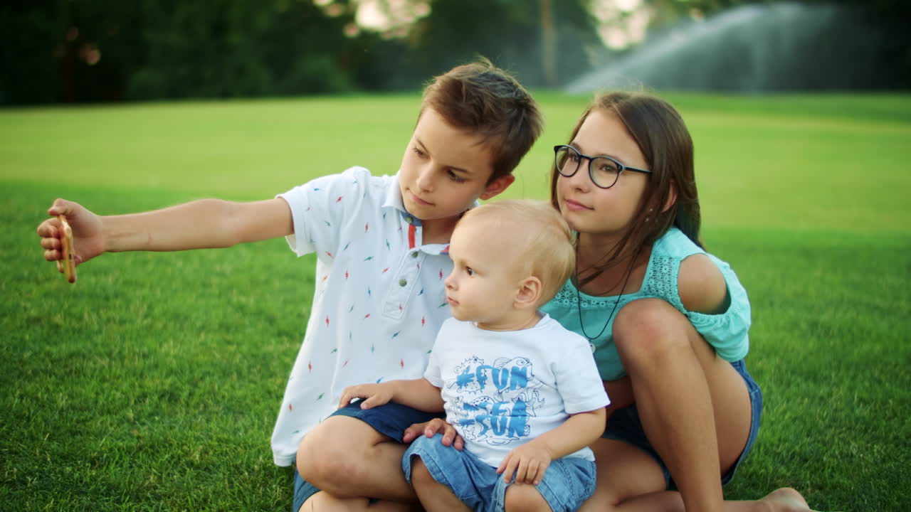 niños y niñas sonrientes sentados en el prado verde. niños tomando selfies en teléfonos inteligentes