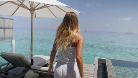 female in overwater bungalow opens window curtains and walks on pool deck, sea view.