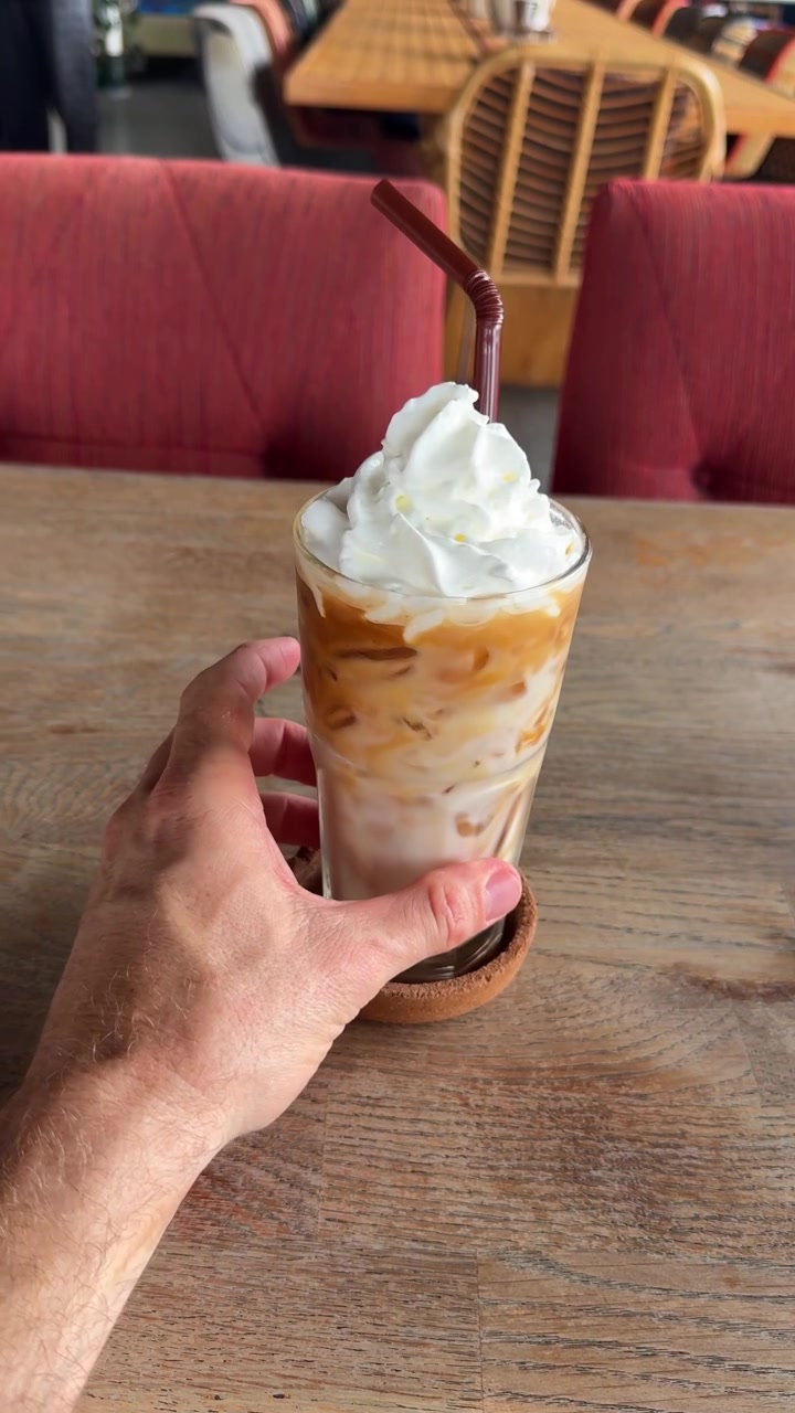 Mano sosteniendo un refrescante café helado cubierto con crema batida dentro de una acogedora cafetería. Perfecto para la cultura del café, bebidas de verano, cafeterías, estilo de vida, hostelería, postres. Primer plano vertical