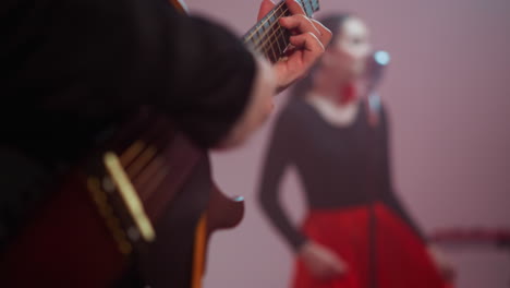 focused capture of a guitarist's hand playing a red guitar, with a blurred female singer in a black and red gown in the background, illustrating a vibrant music scene