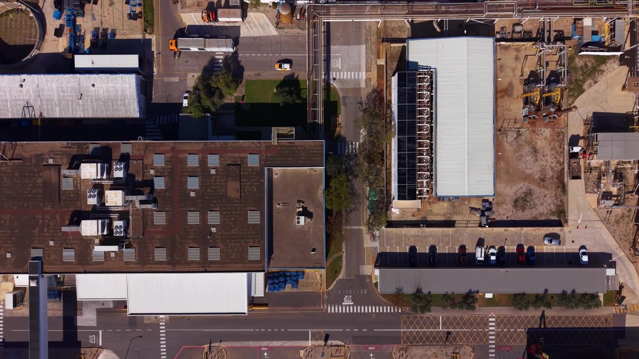 Top-down view of an industrial district showing factory rooftops, warehouse buildings, parking lot with vehicles, road markings and crosswalks in an urban area