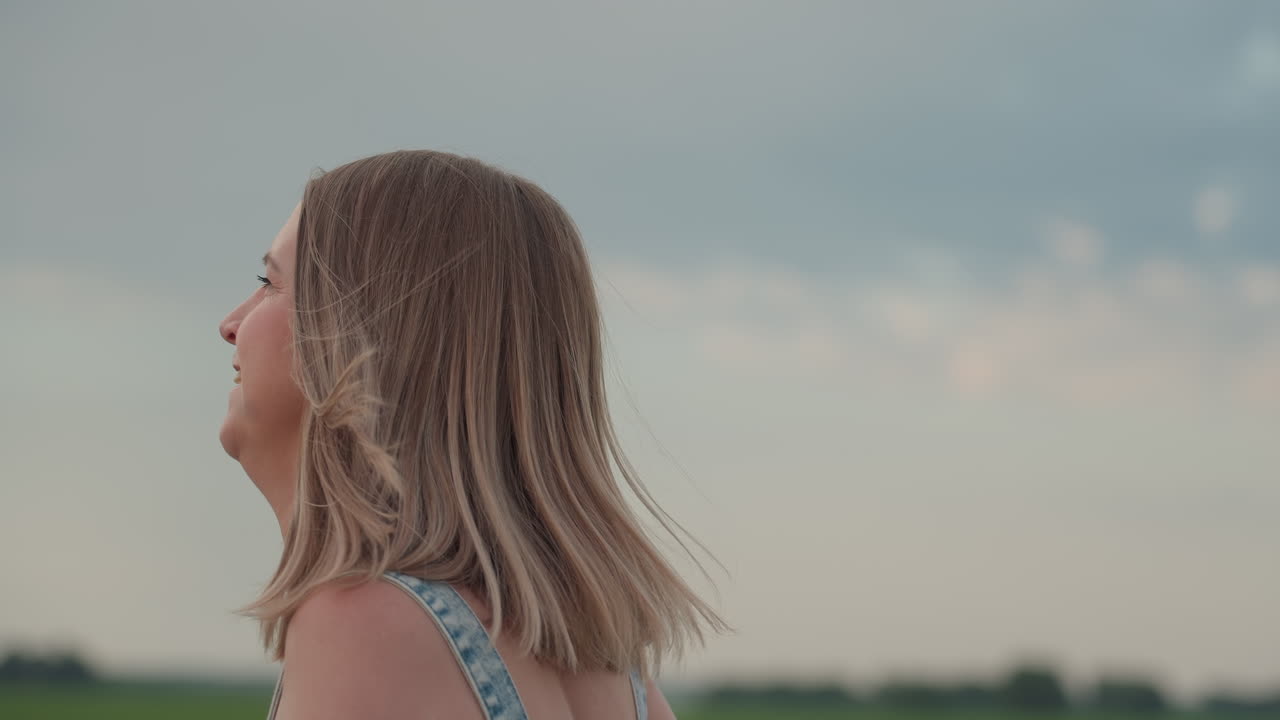 Close up side view of white woman running on asphalt road holding colourful pinwheel fan while wind tosses hair against blurred green field under cloudy sky