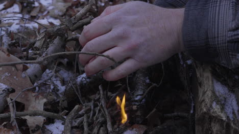 a man uses his hands to shield the tiny flames from the wind as he begins to build a campfire in the winter with leaves and snow on the ground