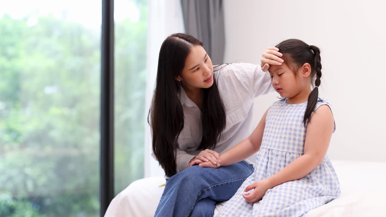 Concerned mother checks daughter’s forehead, offering comfort in softly lit, modern bedroom environment