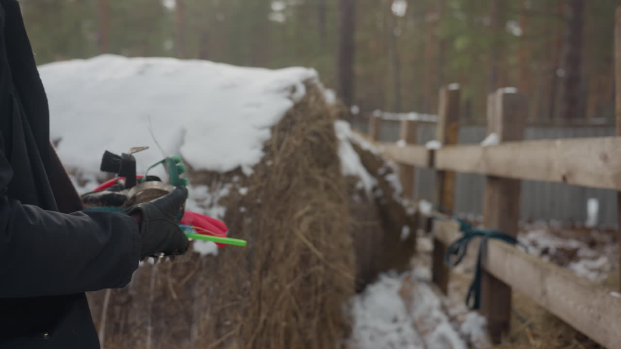 black stablehand preparing grooming brushes outdoors snow-dusted hay bale wooden fence gloved hands holding red and green brushes quiet morning routine tactile focus on tools and motion