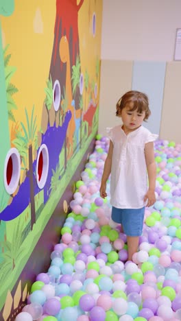 girl playing in a colorful ball pit, throwing balls into holes on a dinosaur-themed wall - vertical slow motion