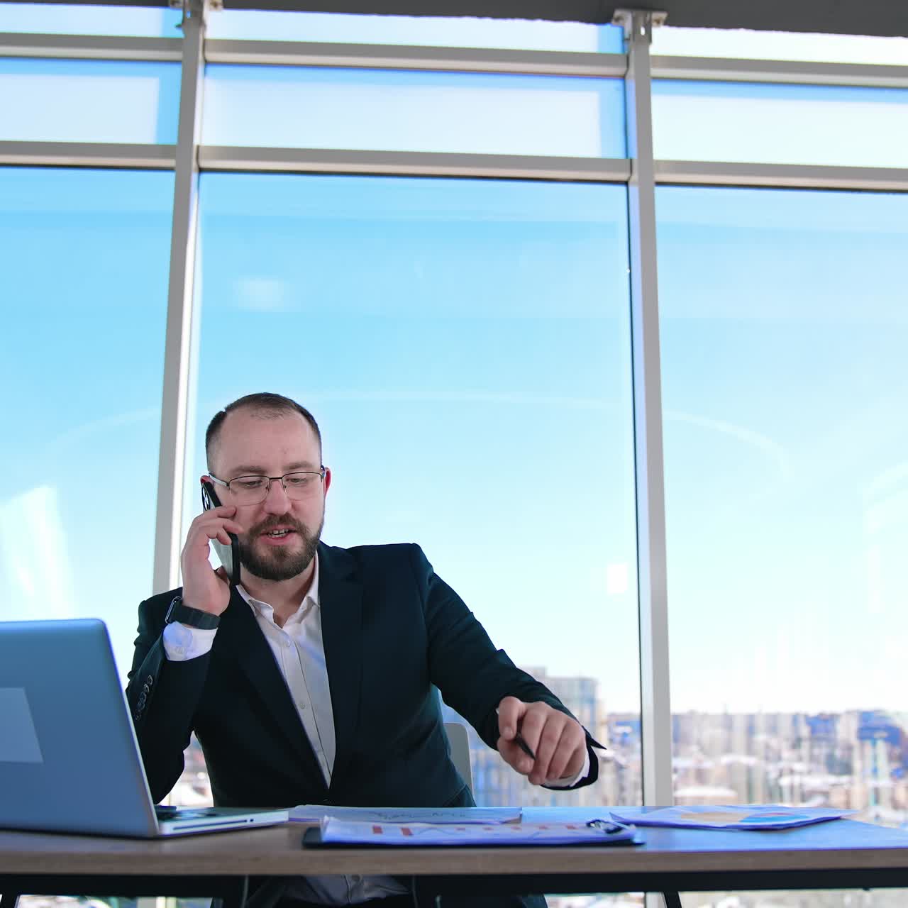 Portrait of entrepreneur at workplace. Confident businessman in glasses talking the phone while sitting in front of a laptop in office