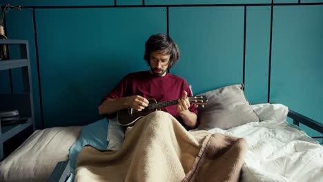 a brunette guy with a beard in a red shirt with glasses plays the guitar lying on a bed with blue walls