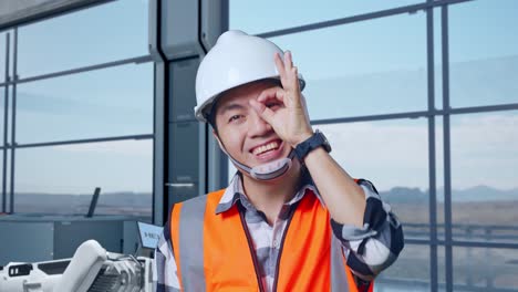 primer plano de un ingeniero asiático con casco de seguridad de pie en una fábrica de alta tecnología, analizando el concepto de la máquina robótica. sonriendo a la cámara y mostrando el signo de la mano ok sobre el ojo