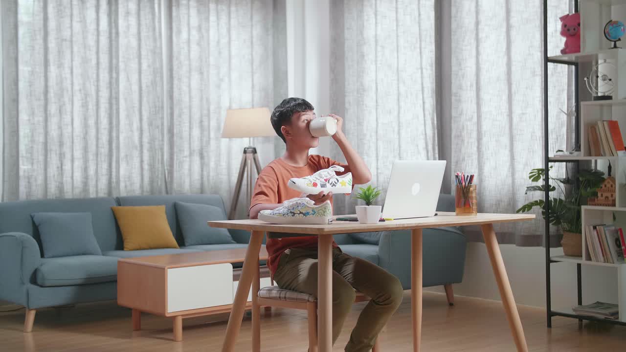 아시아 소년 신발 디자이너가 집에서 노트북에서 일하면서 커피를 마시고 있습니다.