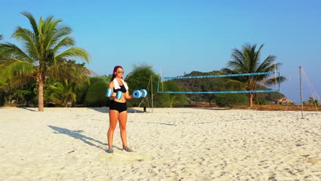 Young-woman-in-sportswear-training-weight-lifting-balance-on-quiet-beach-with-wide-white-sand-on-shore-of-tropical-island-in-Bali