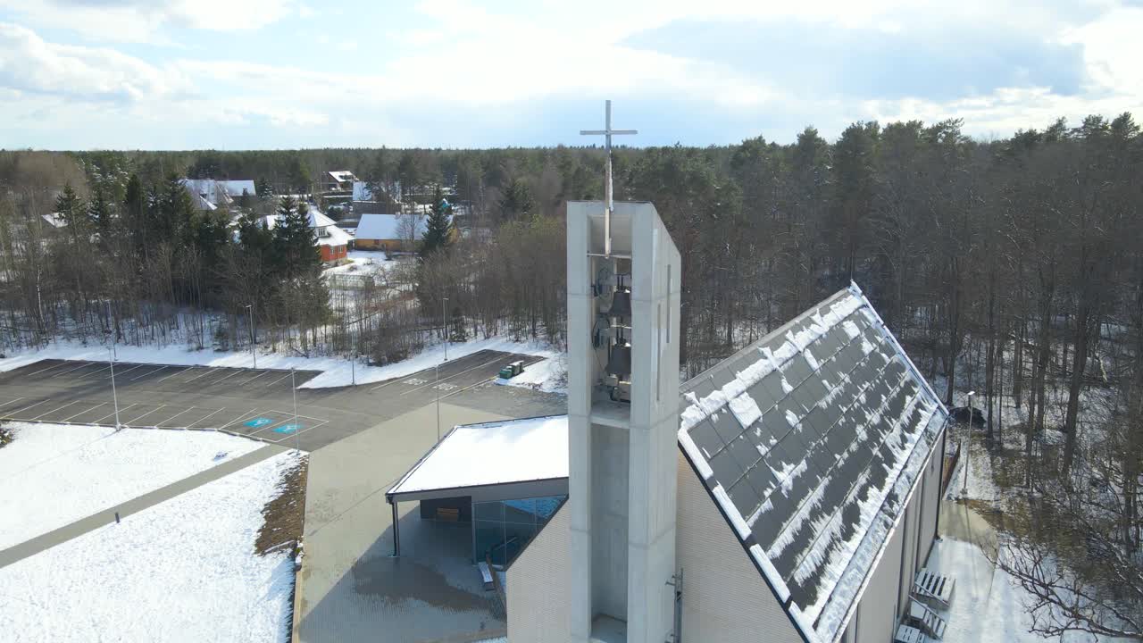 Aerial descending view of modern Lutheran church during sunny snowy day in Saku, Estonia. High angle elevated view of modern religious building with bells and big cross at the top of the church tower.