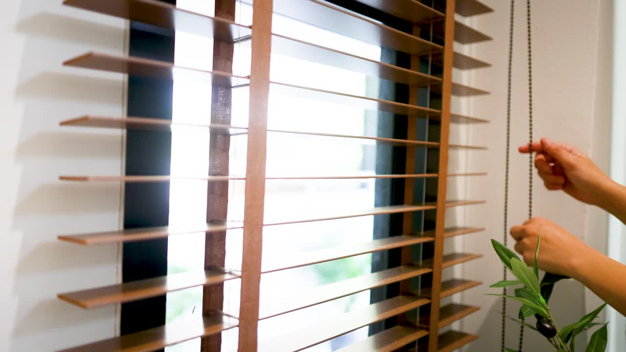 Primer plano de manos tirando de un cordón para abrir persianas de madera, dejando entrar la luz brillante del sol