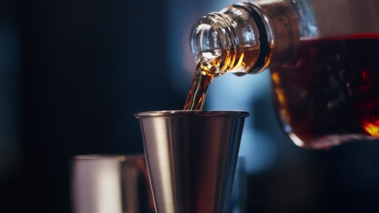 Primo piano di un distillato scuro versato in un misurino di metallo in un cocktail bar con luci soffuse.