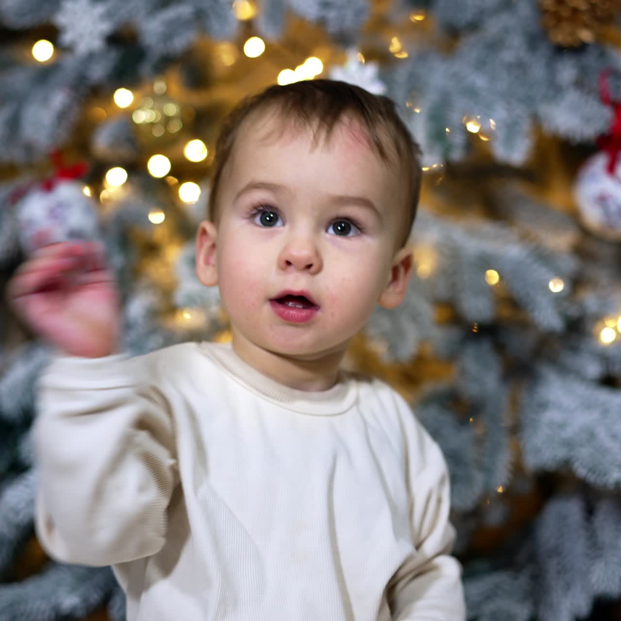 Pretty little kid sits over a decoration for Christmas tree. Adorable child enjoys the toy and waves hands excitedly