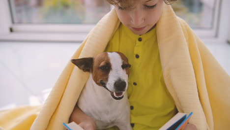 blond boy with curly hair sitting on the floor covered with a blanket next to his dog while reading 1