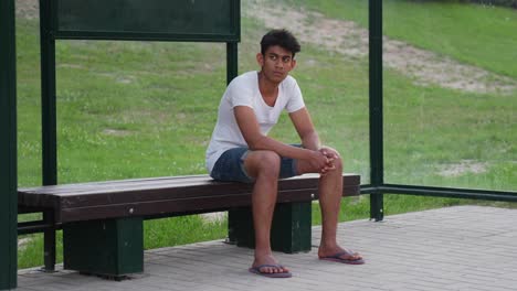 movimiento lento de un joven de sri lanka en ropa básica y zapatillas casualmente sentado y esperando en la parada de autobús cerca de los campos