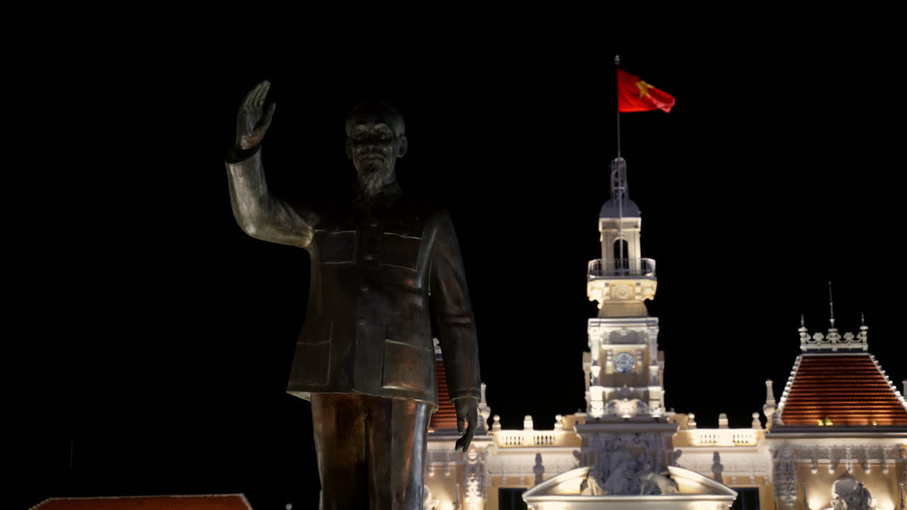 Estátua de Ho Chi Minh iluminada à noite na praça central com edifícios cívicos próximos, estabelecendo foco médio em rack para o prédio do governo