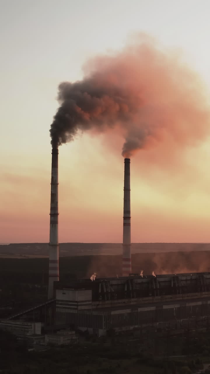 Harmful factory near the river at dusk. Industrial zone with chimneys pouring dark smoke into the air at sunset. Pollution of the environment. Aerial view. Vertical video