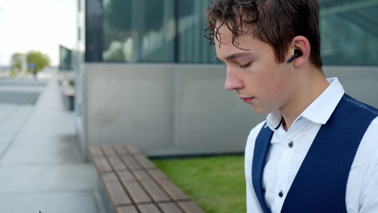 Young Businessman In Suit Wearing Earbuds, Typing On Laptop Keyboard While Sitting On Bench Outside Glass Building. side view, dolly-in shot
