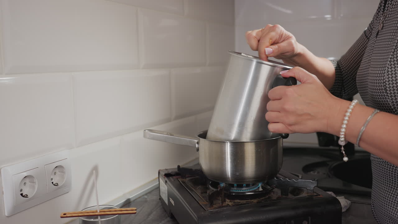 Partial view of woman holding silver container over boiling pot on gas stove while stirring contents, scene includes tiled kitchen wall, nearby candle setup, and electric socket
