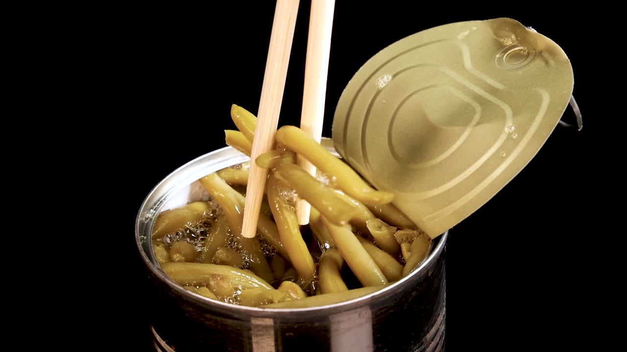 Chopsticks grasp and lift haricot beans from an open can against a black background, with bright, even lighting and a static camera angle