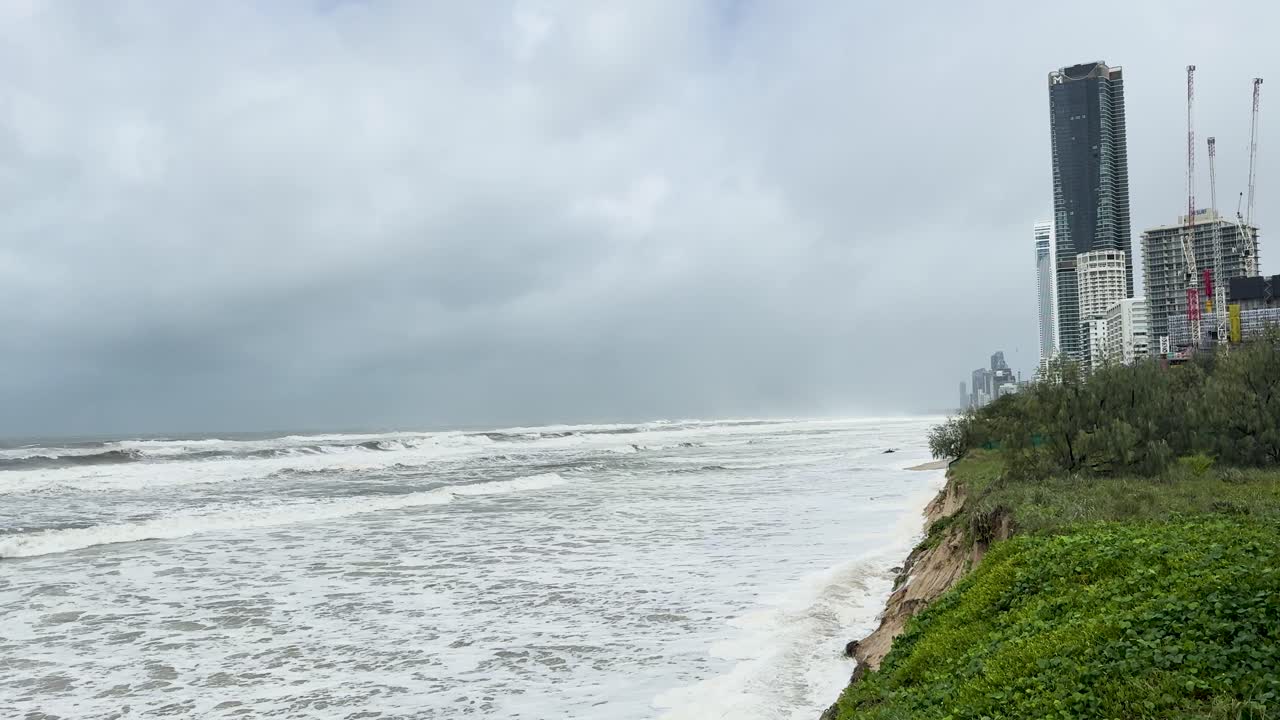 강한 파도가 흐린 바다 풍경으로 구름이 많은 하늘 아래 고층 건물 근처의 해변을 침식합니다.
