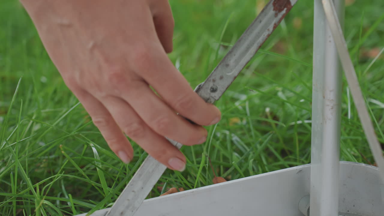 Closeup camping table hinge mechanics, hand adjusts metal bracket and rivet, detailed movement of pivot, signs of wear and rust, focus on maintenance and function, green grass background