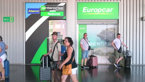 train passengers walk through the frame while others wait outside the car rental companies europcar, alamo, enterprise, and national at alicante train station in alicante, spain.