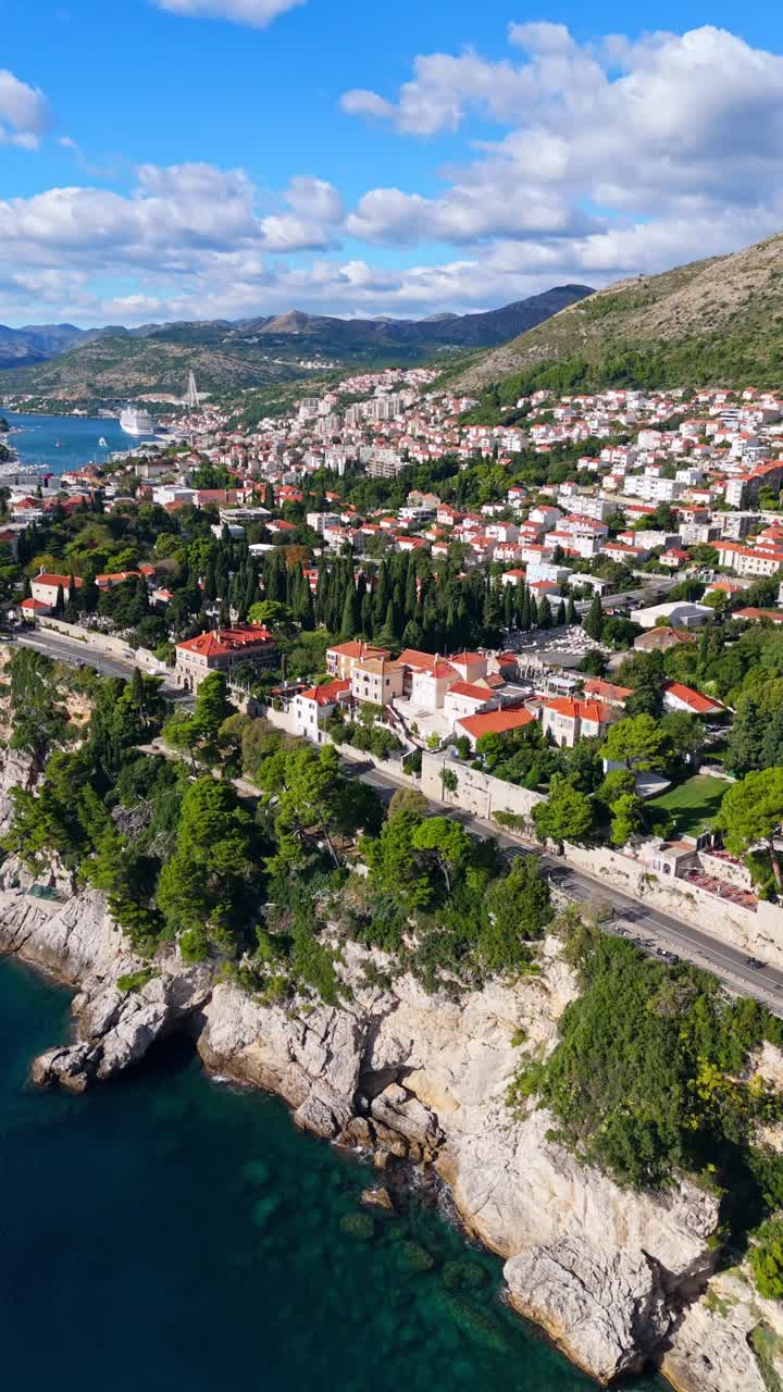 Aerial portrait drone captures Dubrovnik’s cliffside coast, showing terracotta-roofed town buildings, lush green trees, and rugged cliffs bathed in warm sunlight highlighting the dramatic Adriatic Sea