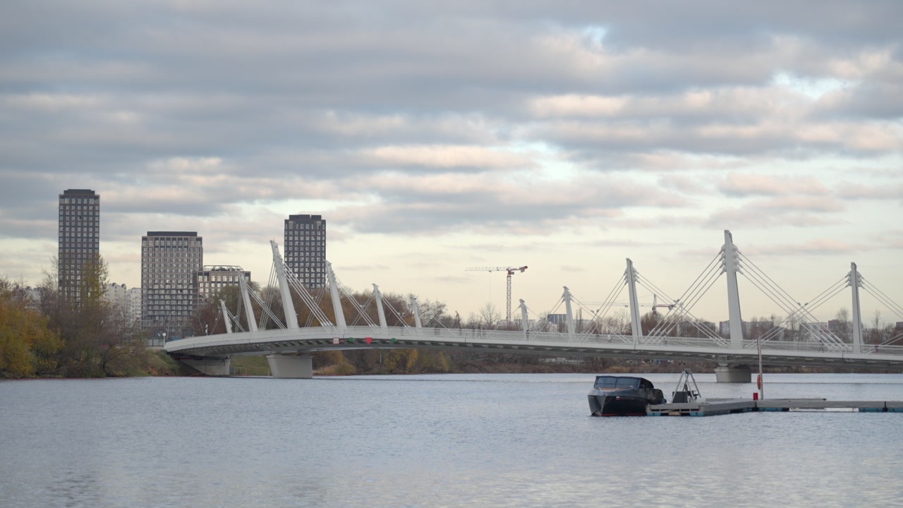 Calm River Waterfront With Bridge, Gray Clouds And Distant Towers, Moored Boat At Small Dock, Gentle Water. Creative