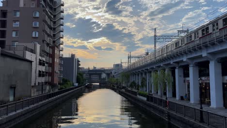 Zug,-Der-Bei-Einem-Wunderschönen-Sonnenuntergang-In-Einer-Städtischen-Umgebung-über-Eine-Kanalbrücke-Fährt