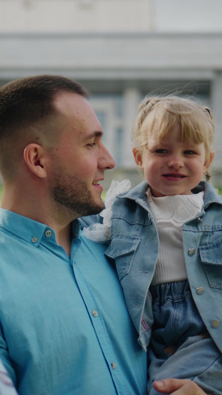 Un uomo porta una bambina allegra all'aperto. Lei indica verso l'alto con espressione gioiosa. Lui guarda in alto con un sorriso dolce. La scena cattura un tenero momento padre-figlia. Ideale per famiglia, genitorialità.