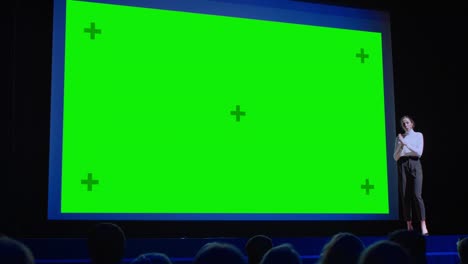 business conference stage: female keynote speaker presents new product to the audience, movie theater shows green screen, mock-up, chroma key. live event on health, science and technology