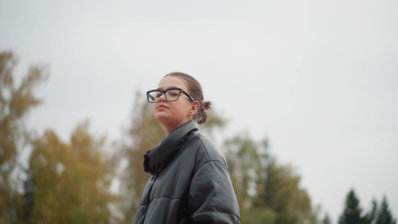 Primer plano de una mujer con gafas y cabello oscuro en un entorno exterior, vistiendo una chaqueta de cuero negra. El sujeto permanece inmóvil bajo una iluminación suave y nublada, mientras el follaje otoñal se difumina en el fondo.