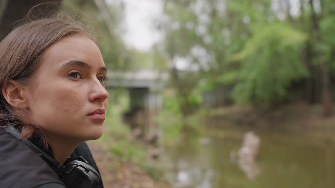 woman by creek in reflective mood, student pauses near muddy river under bridge, soft overcast light, leafy trees and mossy log, profile gaze toward water, cinematic shallow depth of field capturing