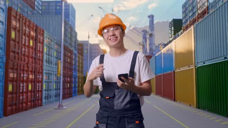 trabajador asiático con gafas de protección y casco de seguridad sonriendo y señalando el teléfono inteligente en su mano mientras está de pie en el almacén del patio de contenedores