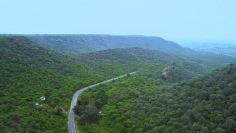 Vista-Aérea-De-Un-Camino-A-Través-De-Un-Bosque-Exuberante-Selva-Verde-Con-Un-Telón-De-Fondo-Montañoso-Durante-El-Monzón-En-Gwalior-Madhya-Pradesh-India