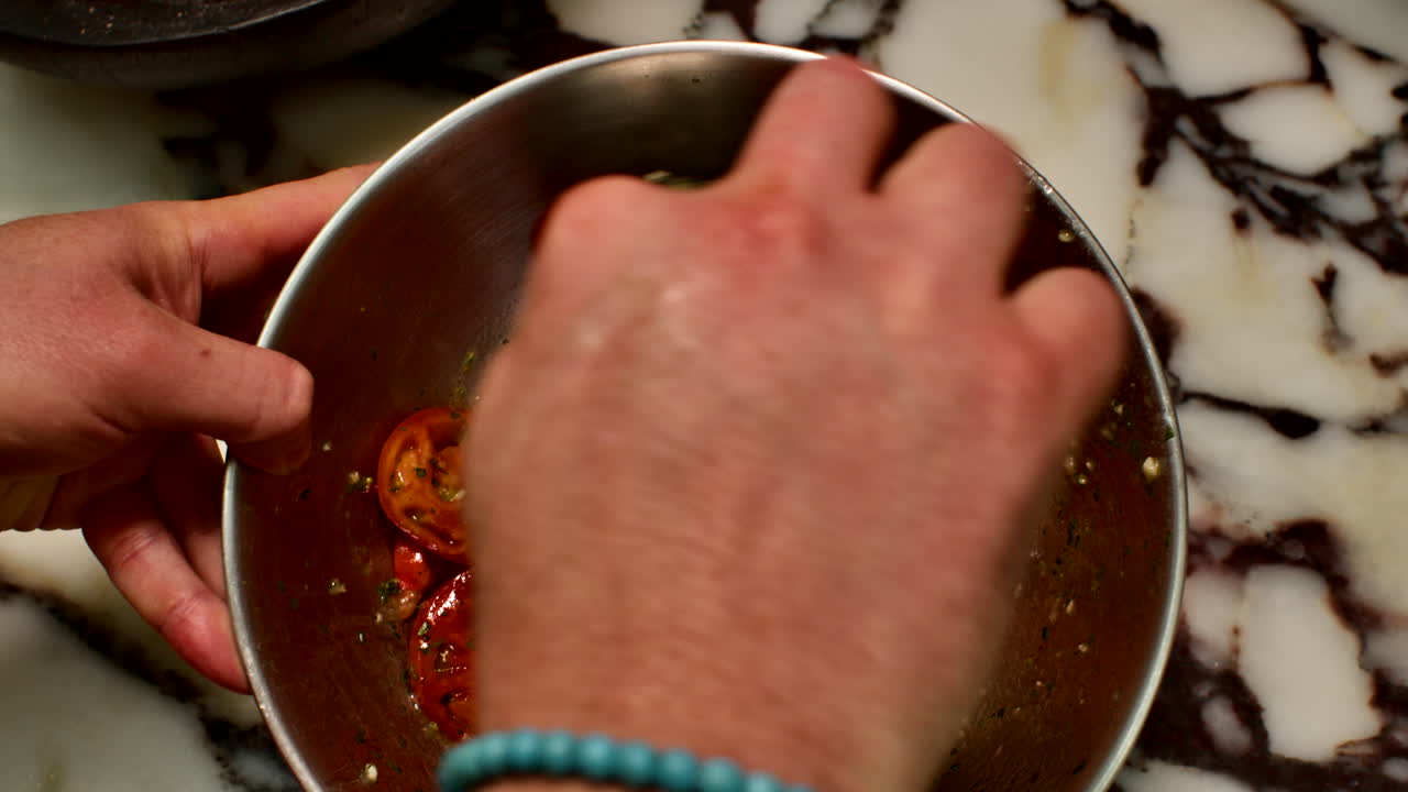 Close-up of a chef's hands tossing roasted tomatoes with fresh herbs in a metal bowl. Professional culinary food styling and preparation in a generic kitchen setting