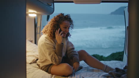 joyful traveller talking cell phone sitting trailer bed on evening ocean coast.