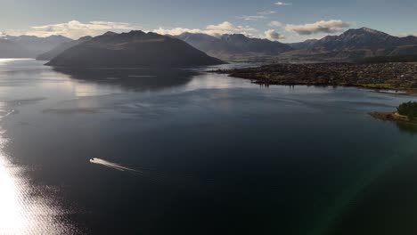Epic aerial view of speedboat on Lake Wanaka with sun reflection on water, revealing town and snow-capped Southern Alps, New Zealand