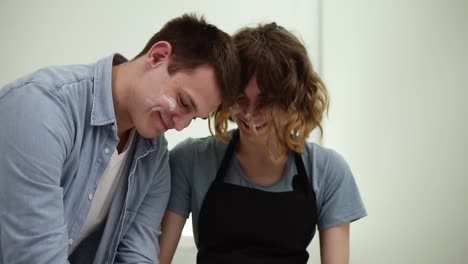 Attractive-caucasian-man-preparing-dough-on-the-white-counter-on-kitchen.-His-girlfriend-helping-him-with-that.-Young-couple-having-fun,-faces-in-flour.-Slowmotion-shot