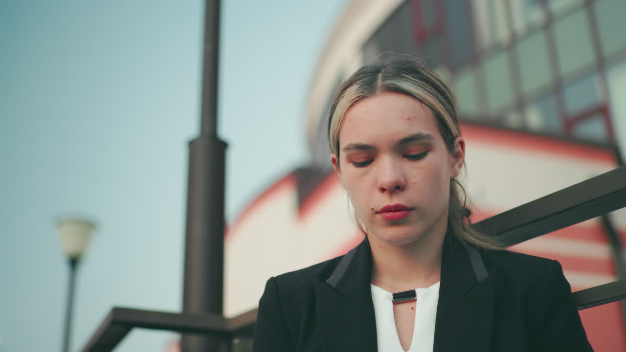 Lady lost in thought in black suit gazes into distance with solemn expression, outdoor urban setting featuring iron pole, modern building with striped design and soft evening lighting in background