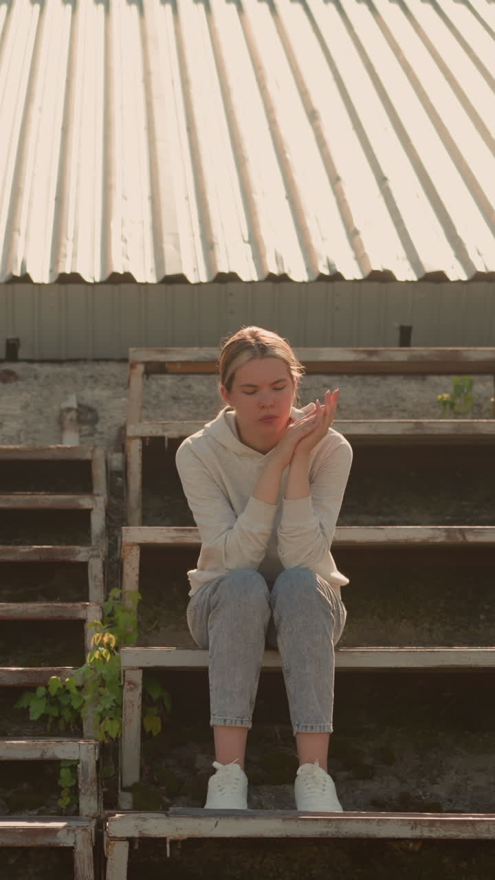 Pensive woman in casual attire sits on rustic stadium bleachers, hands joined together, looking downward thoughtfully, she appears deep in reflection