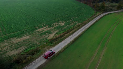 aerial drone forward moving shot over red car driving through road among agriculture fields along green countryside during daytime