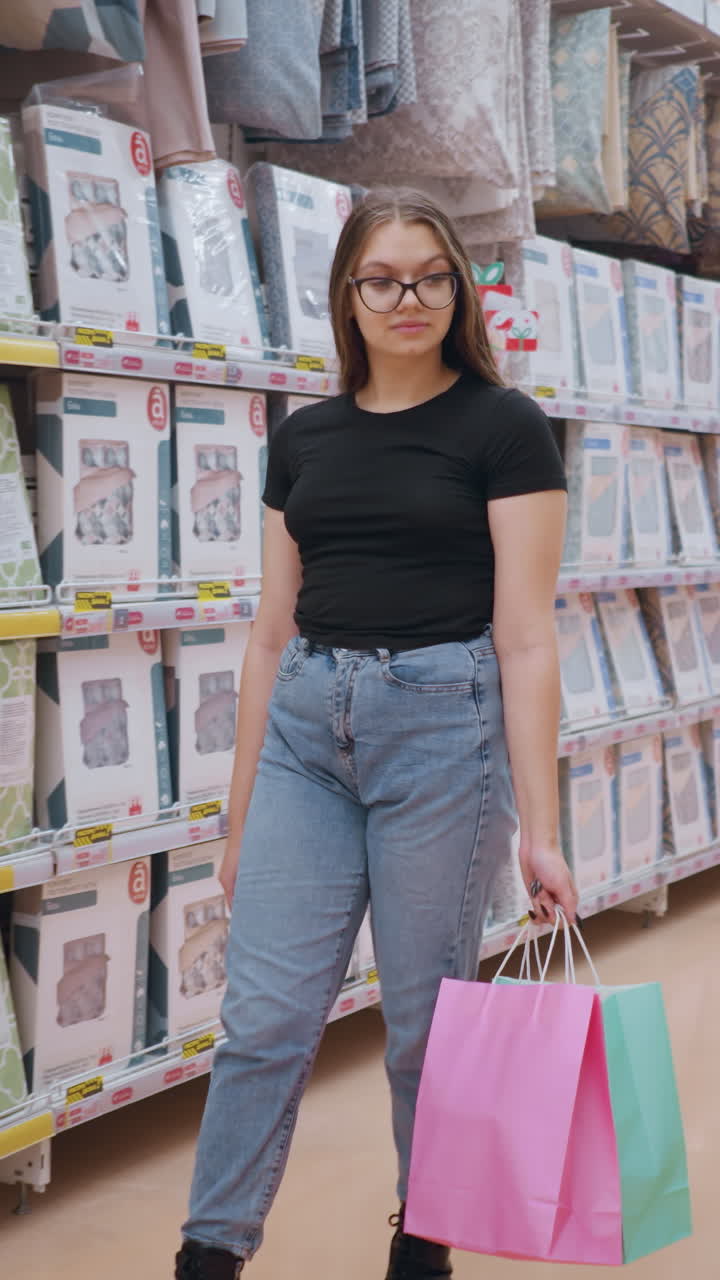 dama de camiseta negra y vaqueros caminando por el pasillo del supermercado con bolsas de compras, centrándose en los productos exhibidos en los estantes, ropa de cama y textiles para el hogar bien dispuestos en un ambiente de venta al por menor brillante